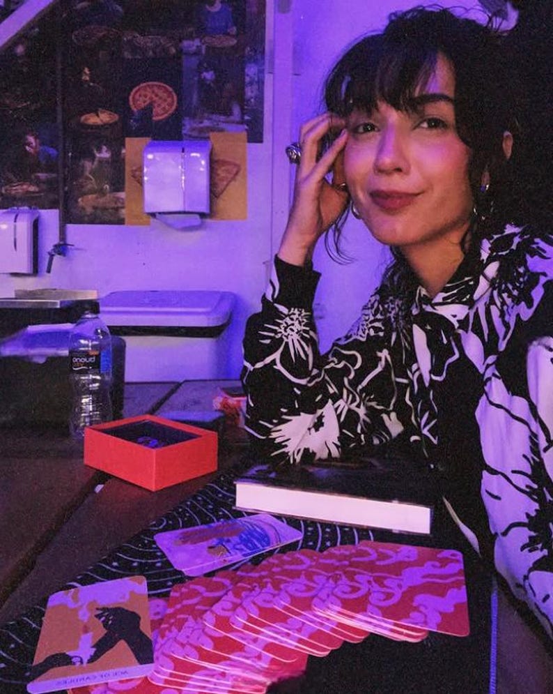 May include: A woman wearing a black and white floral print top, seated at a table with tarot cards. A red box and a book are on the table. The background includes pizza-themed posters and a water bottle.