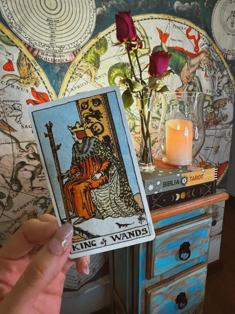 May include: A tarot card depicting the King of Wands, held in front of a vintage map. The card features an illustration of a king in orange and gold robes. A candle in a glass holder and a vase of roses are also visible.