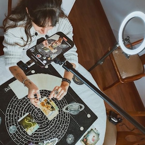 May include: A person is seen from above, arranging tarot cards on a black mat with a celestial design. The mat features concentric circles, planets, and moons. A smartphone is mounted above the cards, and a ring light illuminates the scene.
