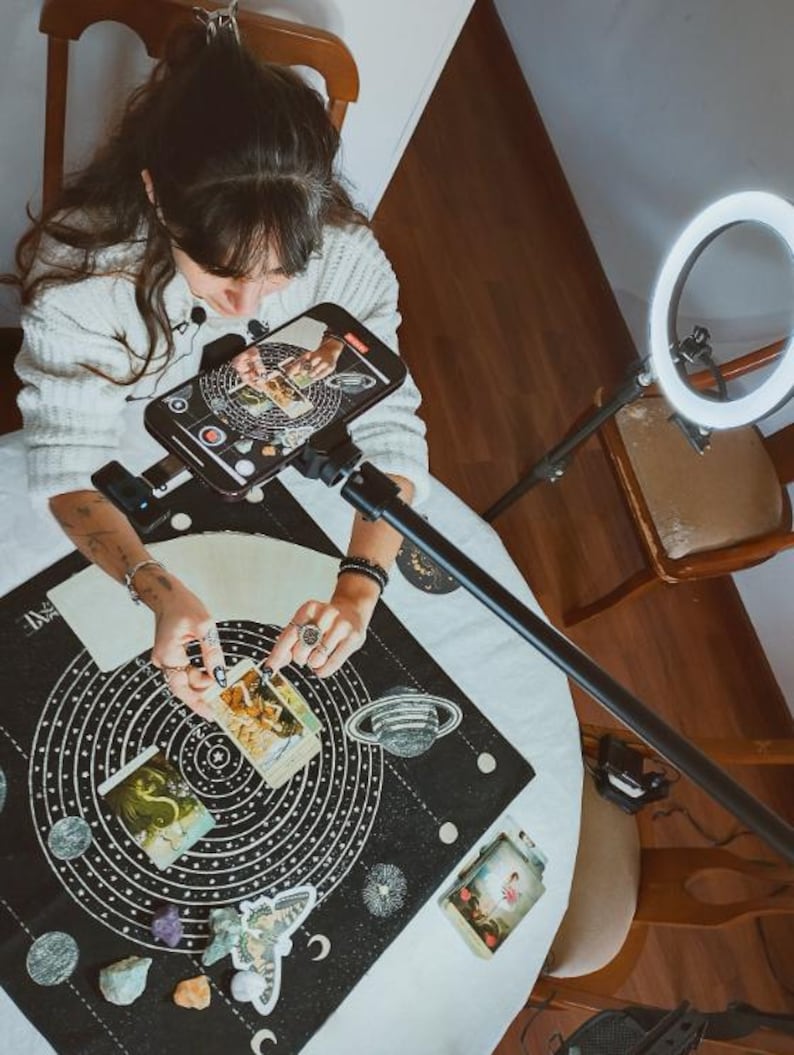 May include: A person is seen from above, arranging tarot cards on a black mat with a celestial design. The mat features concentric circles, planets, and moons. A smartphone is mounted above the cards, and a ring light illuminates the scene.