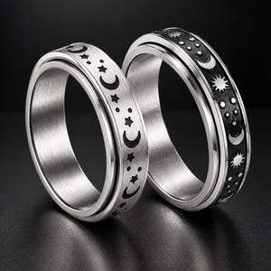 Op de afbeelding: Twee zilverkleurige metalen spinnerringen met een hemels motief. De ringen zijn gegraveerd met zwart ingevulde patronen van maansikkels, zonnen en sterren. De ringen hebben een gepolijste afwerking op de randen en een geborstelde textuur.
