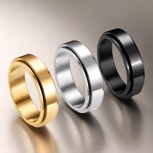 Könnte beinhalten: Drei Spinner-Ringe in einer Reihe angeordnet, bestehend aus einem goldfarbenen, einem silberfarbenen und einem schwarzen Ring. Jeder Ring hat ein glattes, poliertes Außenband, das sich um ein zentrales Innenband dreht, mit metallischem Glanz.
