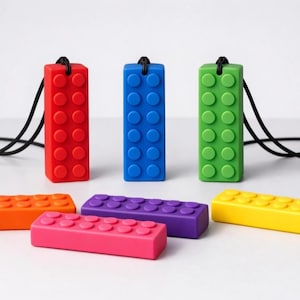 May include: A selection of colourful brick-shaped pendants and blocks. The pendants are red, blue, and green, each with a black cord. The blocks are orange, purple, pink, and yellow. The items are arranged on a white surface.