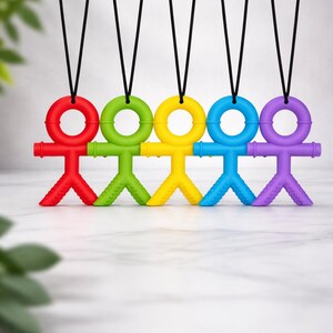 Peut inclure: Une rangée de jouets de dentition colorés en rouge, vert, jaune, bleu et violet. Chaque jouet a la forme d'un bonhomme avec une tête circulaire et est suspendu par un cordon noir.