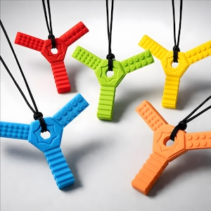 Puede incluir: Cinco collares de dentición de silicona de colores rojo, verde, amarillo, azul y naranja. Cada collar presenta un colgante de tres puntas con un diseño central en forma de estrella y una superficie texturizada, suspendido de un cordón negro.