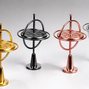 May include: Four metal gyroscopes standing on conical bases, arranged in a row. Each gyroscope features a central vertical axis, a horizontal disc, and a surrounding ring. The colors are gold, black, rose gold, and silver, all with a polished finish.