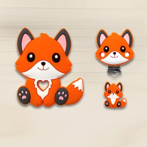 Pode incluir: Três itens em forma de raposa laranja. O maior é uma raposa com um recorte em forma de coração, patas pretas e peito branco. O segundo é uma raposa com um clipe de metal. O menor é uma raposa com as patas juntas.
