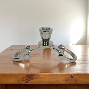 May include: A silver metal candle holder with a central cup for the candle and three curved arms. The candle holder is on a wooden surface. The design is modern and minimalist. The measurements are in centimetres.