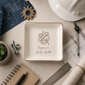 Pode incluir: Uma bandeja quadrada de cerâmica branca com borda dourada, com um design de tema de engenharia e o nome "Linda Smith". O design inclui engrenagens, uma régua e um compasso. Outros itens incluem um capacete, plantas e uma suculenta.