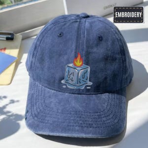 May include: A blue baseball cap featuring an embroidered design of a melting ice cube with a flame. The word "EMBROIDERY" is on a small rectangular patch.