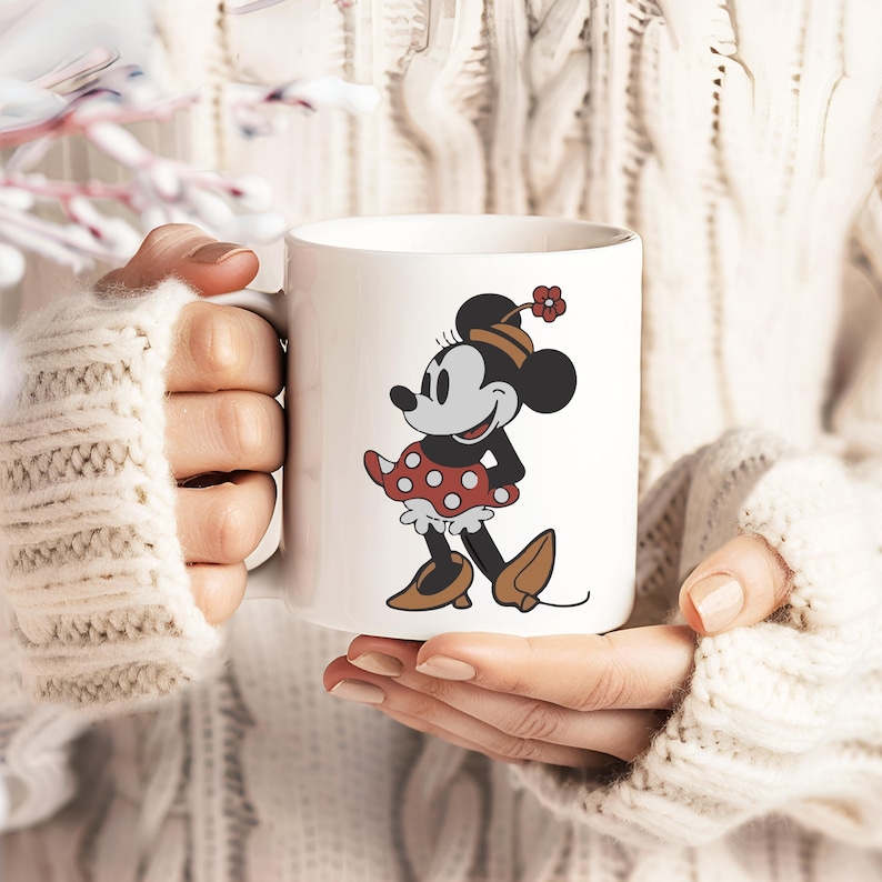 Puede incluir: Taza de cer&aacute;mica blanca con un personaje de dibujos animados cl&aacute;sico con un vestido de lunares rojos, una parte superior negra y zapatos marrones. El personaje lleva un sombrero con una flor roja. La taza la sostiene una persona que lleva un su&eacute;ter blanco.