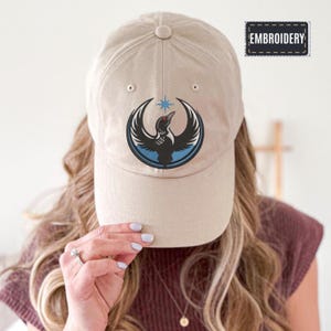 May include: A tan baseball cap with a black and blue embroidered design of a bird with outstretched wings. The cap has a curved brim and a button on top. The word "EMBROIDERY" is printed on a small black rectangle.
