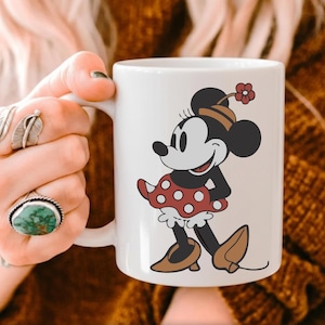 Puede incluir: Taza de cer&aacute;mica blanca con una ilustraci&oacute;n de Minnie Mouse. Minnie lleva su atuendo cl&aacute;sico: una falda roja de lunares, zapatos marrones y un sombrero con una flor. La taza es sostenida por una persona con anillos.