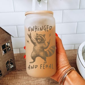 以下が含まれることがあります： 竹製の蓋が付いた、すりガラスのタンブラー。 「UNHINGED AND FERAL」の文字とアライグマのグラフィックが特徴です。 アライグマは、ロックンロールのジェスチャーで腕を上げて描かれています。 タンブラーには明るい色の飲み物が入り、ストローが付いています。