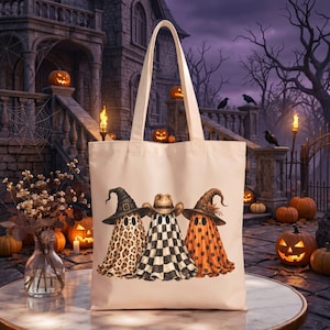 Puede incluir: Un bolso de tela color crema con un estampado gráfico de tres fantasmas con sombreros. El fantasma de la izquierda tiene un patrón de leopardo, el del centro un patrón de cuadros blanco y negro, y el de la derecha tiene lunares naranjas.