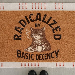 Könnte beinhalten: Eine rechteckige Fußmatte mit braunem Hintergrund und weißem Rand mit roten Punkten. Die Matte zeigt eine Illustration einer grummeligen Katze, die eine Tasse und ein Buch hält, mit dem Text "RADICALIZED BY BASIC DECENCY" in Schwarz.