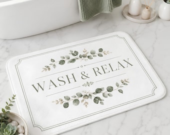 Puede incluir: Una alfombrilla de baño rectangular blanca con un borde gris fino, ilustraciones de hojas de eucalipto en acuarela verde y el texto "WASH & RELAX" impreso en una fuente serif gris oscuro centrada entre los diseños botánicos.