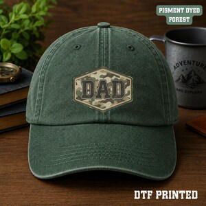 Könnte beinhalten: Eine pigmentgefärbte Baseballkappe in Waldgrün mit einem zentralen sechseckigen Aufnäher in Camouflage-Optik und dem Wort DAD, das in einer dunklen, fetten Blockschrift aufgedruckt ist. Die Kappe hat eine strukturierte Krone und einen gebogenen Schirm.