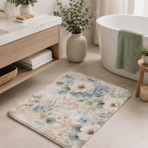 May include: A rectangular bathroom rug featuring a watercolor-style floral pattern with large white flowers, blue hydrangea clusters, and soft green eucalyptus leaves on a light beige background with rounded corners and a finished edge.