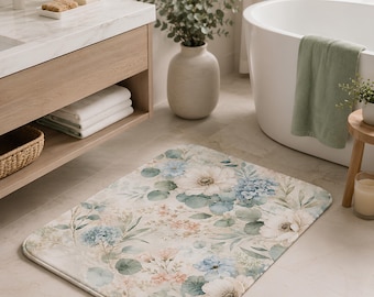 Puede incluir: Una alfombra de baño rectangular con un estampado floral estilo acuarela que presenta grandes flores blancas, racimos de hortensias azules y hojas de eucalipto verde suave sobre un fondo beige claro, con esquinas redondeadas y bordes rematados.