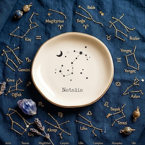 May include: A cream-colored plate with a gold rim, featuring a constellation design and the name "Natalia." Surrounding the plate are gold-colored constellation charms and zodiac symbols on a dark blue fabric. Additional decorative elements include crystals and dried lavender.