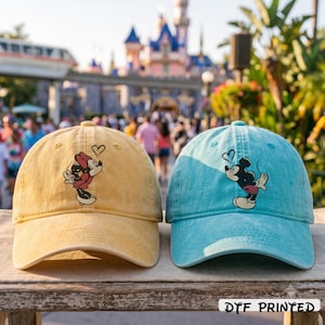 K&ouml;nnte beinhalten: Eine gelbe Baseballkappe mit einer Grafik von Minnie Maus, die ein Herz bl&auml;st, und eine hellblaue Baseballkappe mit einer Grafik von Mickey Maus, die ein Herz bl&auml;st. Beide Kappen haben ein verwaschenes Design und den Text DTF PRINTED unten.