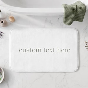 Può includere: Un tappetino da bagno rettangolare bianco con angoli arrotondati che presenta il testo custom text here stampato in un carattere serif grigio centrato. Il tappetino ha una consistenza morbida e soffice, progettato per il pavimento del bagno.