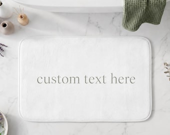 Puede incluir: Una alfombrilla de baño rectangular blanca con esquinas redondeadas que presenta el texto custom text here impreso en una fuente serif gris centrada. La alfombrilla tiene una textura suave y afelpada, diseñada para el suelo del baño.