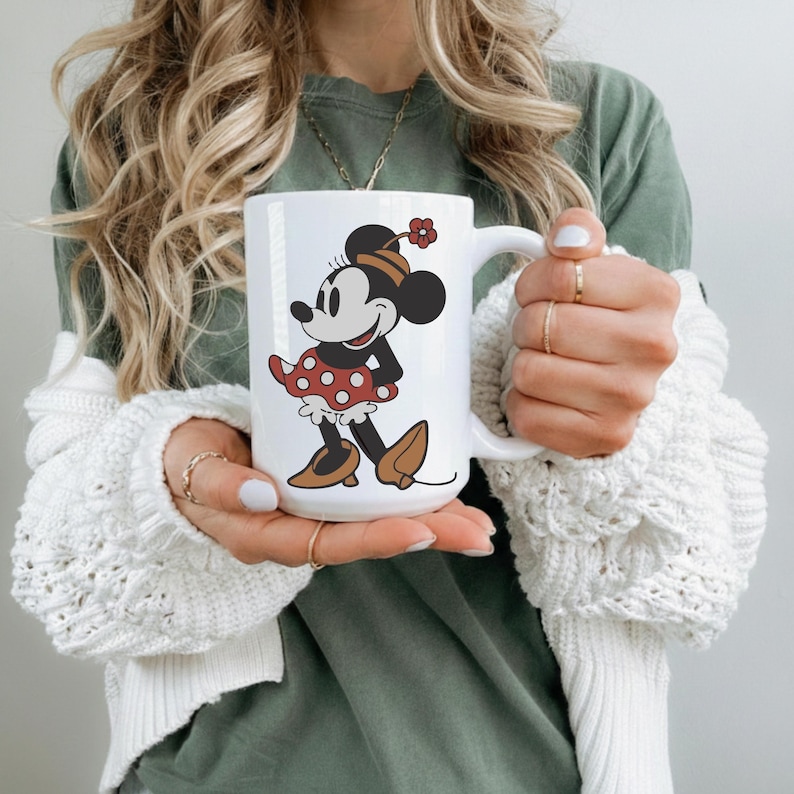Puede incluir: Taza de cer&aacute;mica blanca con una imagen de dibujos animados de Minnie Mouse con un vestido de lunares rojos y zapatos marrones. La taza tiene un asa grande. El dise&ntilde;o incluye una flor roja en el sombrero de Minnie.