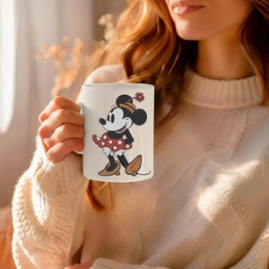 Puede incluir: Taza de cer&aacute;mica blanca con una ilustraci&oacute;n de dibujos animados de Minnie Mouse. Minnie aparece con un vestido rojo de lunares, zapatos marrones y un sombrero a juego. La taza la sostiene una persona que lleva un su&eacute;ter color crema.