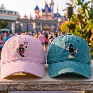 K&ouml;nnte beinhalten: Eine hellrosa Baseballkappe mit einer Grafik von Minnie Maus, die ein Herz bl&auml;st, und eine blaugr&uuml;ne Baseballkappe mit einer Grafik von Mickey Maus, die ein Herz bl&auml;st. Beide Kappen haben einen gebogenen Schirm, eine strukturierte Krone und Stickereien.