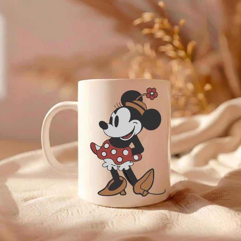 Puede incluir: Una taza de cer&aacute;mica color crema con una ilustraci&oacute;n de dibujos animados de Minnie Mouse. Minnie est&aacute; representada con un vestido de lunares rojos, zapatos marrones y un sombrero a juego con una flor. La taza tiene un asa blanca y est&aacute; sobre un fondo suave y neutro.