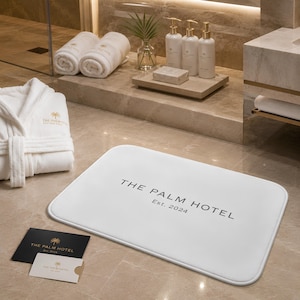 Puede incluir: Una alfombrilla de ba&ntilde;o rectangular blanca con el texto negro THE PALM HOTEL Est. 2024. El conjunto incluye un albornoz blanco con un logo dorado, dos toallas blancas enrolladas, tres botellas con dosificador, jab&oacute;n y dos tarjetas con palmeras.