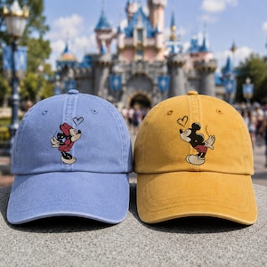 K&ouml;nnte beinhalten: Eine blaue Baseballkappe mit einer Grafik von Minnie Maus, die ein Herz bl&auml;st, und eine gelbe Baseballkappe mit einer Grafik von Mickey Maus, die ein Herz bl&auml;st. Beide Kappen haben eine strukturierte Krone, einen gebogenen Schirm und ein mattes Finish.