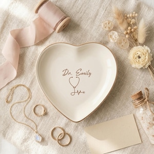 May include: A heart-shaped, off-white ceramic trinket dish with a gold rim. The dish is inscribed with "Dr. Emily" and a stethoscope graphic. Gold jewelry and a spool of pink ribbon are arranged around the dish, suggesting a gift or decorative theme.