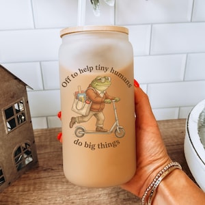May include: Frosted glass tumbler with a bamboo lid and a straw. The tumbler features a whimsical illustration of a frog riding a scooter with the text "Off to help tiny humans do big things."