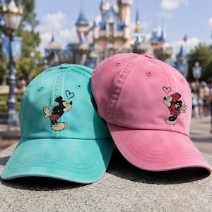 K&ouml;nnte beinhalten: Eine t&uuml;rkisfarbene Baseballkappe mit einer Mickey-Maus-Grafik und eine rosafarbene Baseballkappe mit einer Minnie-Maus-Grafik. Beide Kappen haben ein kleines Herzsymbol &uuml;ber der Figur sowie einen gebogenen Schirm und eine strukturierte Krone.