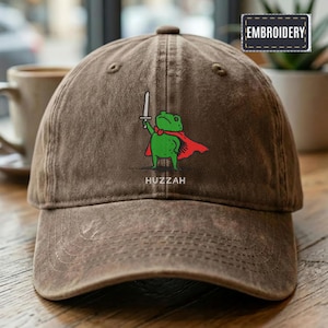 May include: Brown baseball cap with an embroidered cartoon frog wearing a red cape and holding a sword. The frog has a green body and the word "Huzzah" is printed below the frog.