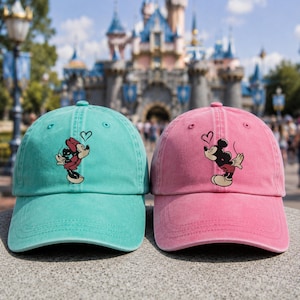 K&ouml;nnte beinhalten: Zwei Baseballkappen, eine in T&uuml;rkis und eine in Pink, mit jeweils einer Grafik einer Zeichentrickmaus, die einen Kuss mit einem kleinen Herzumriss dar&uuml;ber sendet. Die Kappen haben eine strukturierte Krone, einen gebogenen Schirm und &Ouml;sen.