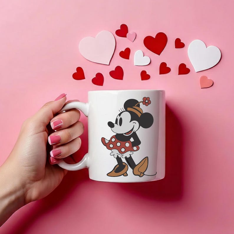 Puede incluir: Taza de cer&aacute;mica blanca con un dise&ntilde;o de Minnie Mouse, con un vestido de lunares rojos y zapatos marrones. La taza est&aacute; sobre un fondo rosa con decoraciones de coraz&oacute;n rojas y blancas. Un art&iacute;culo tem&aacute;tico de Disney.