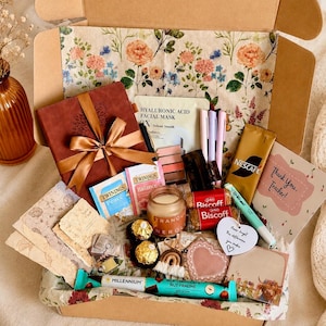 May include: A gift box containing a brown notebook with a ribbon, Twinings tea bags, a candle, Biscoff cookies, a Nescafé stick, a Millennium chocolate bar, a hyaluronic acid facial mask, pens, a heart-shaped tag, and a card reading Thank You, Teacher!
