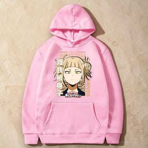 May include: A light pink hooded sweatshirt featuring a graphic print of the anime character Himiko Toga with blonde hair and yellow eyes. The design includes Japanese text, the name Himiko Toga, the word Knives, and the My Hero Academia logo.