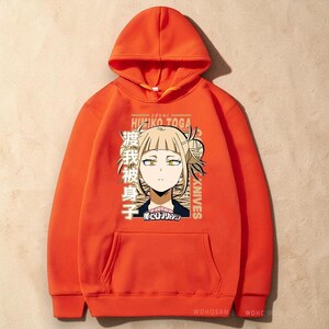 May include: A bright orange hooded sweatshirt featuring a graphic print of the anime character Himiko Toga. The design includes the character's face, Japanese text, the name Himiko Toga, and the My Hero Academia logo centered on the front chest area.