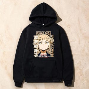 May include: A black hooded sweatshirt featuring a graphic print of the anime character Himiko Toga. The design includes the text Himiko Toga, Knives, and Japanese characters in a tan color palette, along with a small logo at the bottom center.