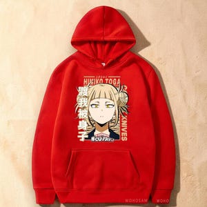 May include: A bright red hooded sweatshirt featuring a graphic print of the anime character Himiko Toga with blonde hair and yellow eyes. The design includes the text Himiko Toga, Knives, and Japanese characters, along with the My Hero Academia logo.