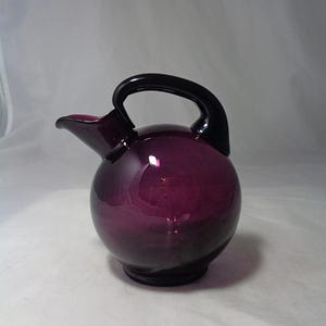 May include: A deep purple glass pitcher with a spherical body, a short pouring spout, and a dark, curved handle attached to the top. The glass is translucent, allowing light to pass through the rounded vessel, which sits on a small, circular base.