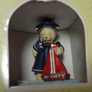 May include: A small, tan teddy bear figurine dressed in a navy sailor suit and hat, holding a red sailboat with the text "USS MUFFY." The figurine is displayed inside a decorative archway.