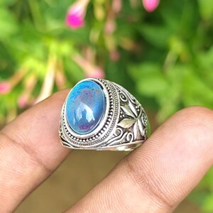 May include: A silver ring featuring an oval-shaped, iridescent blue and purple cabochon stone set in a decorative metal band with intricate floral and leaf-like engravings along the sides and a beaded border surrounding the central gemstone.