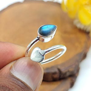 Könnte beinhalten: Silberfarbener Ring mit einem tropfenförmigen, blauen Edelstein. Das Design des Rings weist ein herzförmiges Detail und einen Wickelstil auf. Der Edelstein ist etwa 1,3 cm lang.