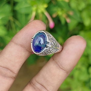 Puede incluir: Un anillo de metal plateado con una piedra cabujón ovalada de color azul profundo en el centro. La banda del anillo presenta un intrincado patrón en relieve que representa alas de libélula a ambos lados del engaste de la piedra central.
